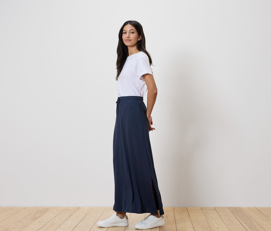 Femme debout de profil, portant un t-shirt blanc, une longue jupe bleu marine et des baskets blanches.