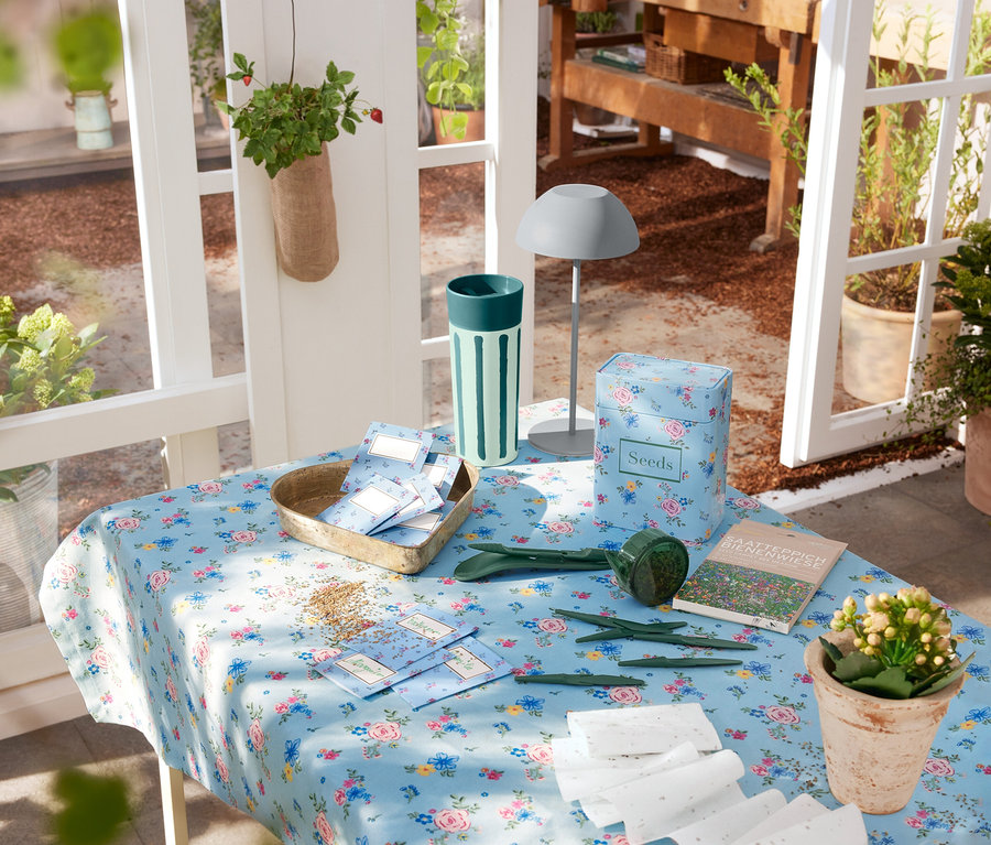 Une lampe de table LED gris аккумуляторная est posée sur une table recouverte d'une nappe à fleurs, à côté d'outils de jardinage et de graines.