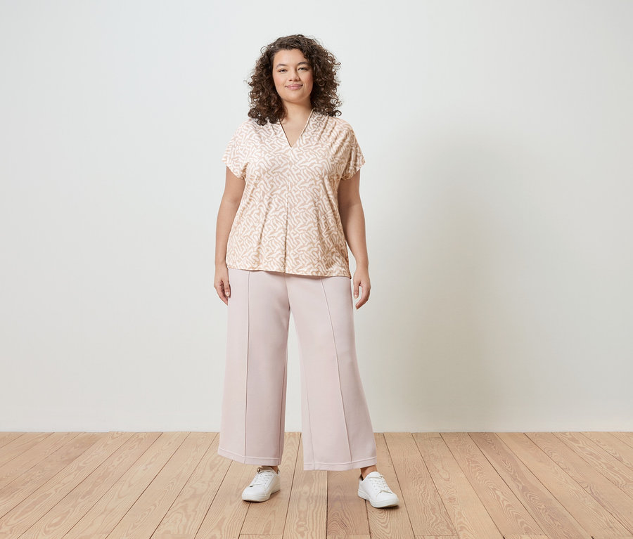 Une femme aux cheveux bouclés porte un chemisier à motifs rose clair, une culotte rose et des baskets blanches devant un mur blanc.