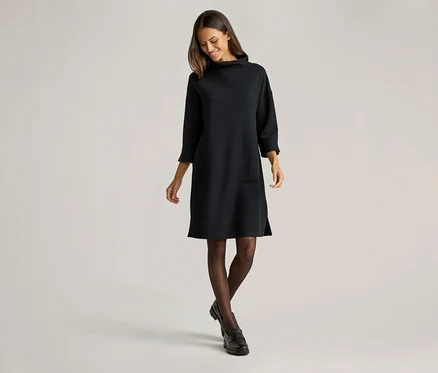 Femme souriante portant une robe noire en maille à col roulé arrivant aux genoux, des collants foncés et des chaussures noires sur fond clair.