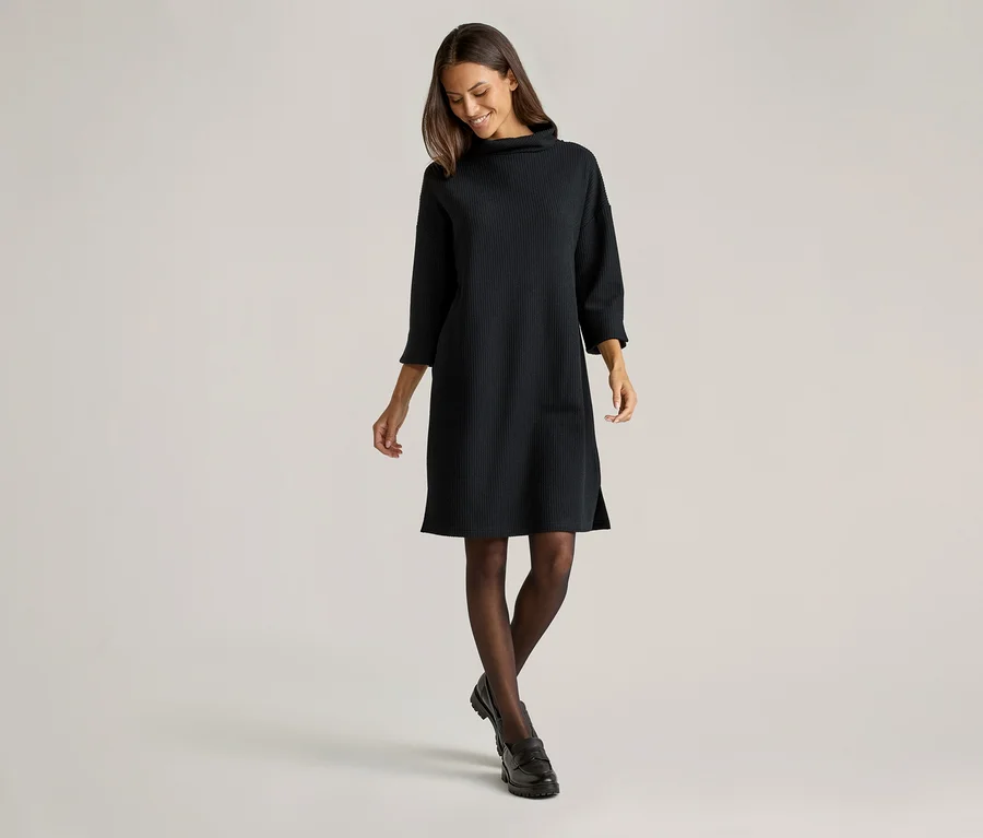 Femme souriante portant une robe noire en maille à col roulé arrivant aux genoux, des collants foncés et des chaussures noires sur fond clair.