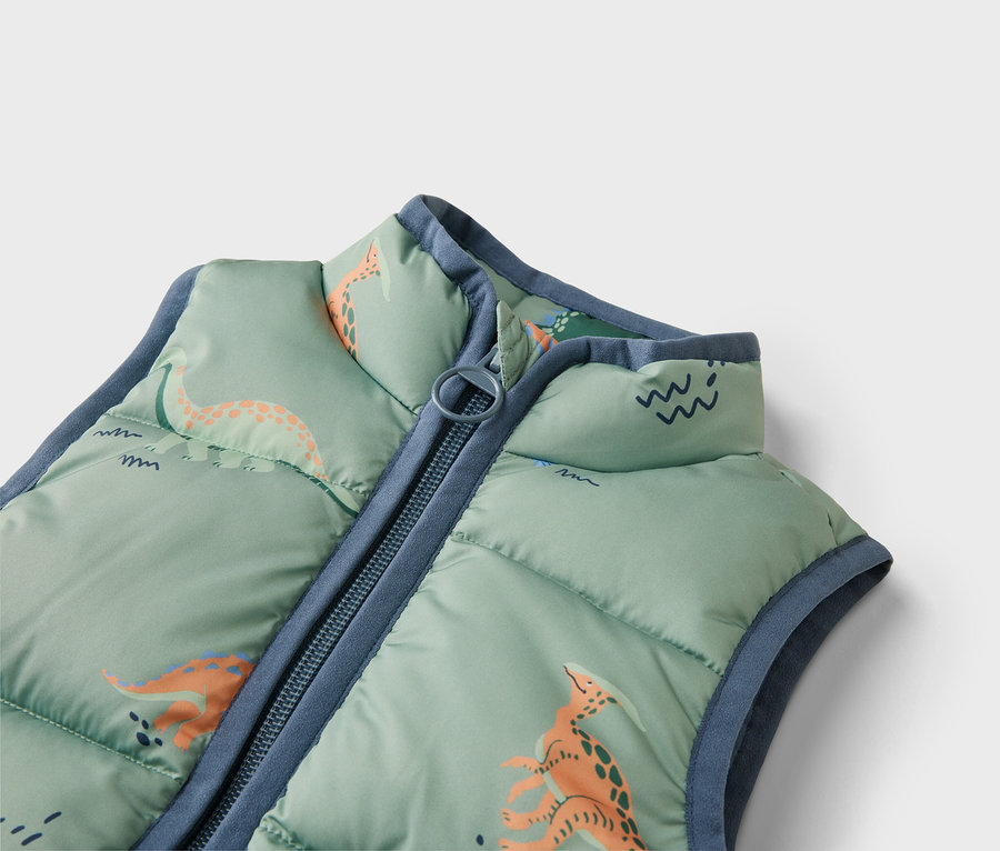 Gros plan d'un gilet matelassé vert clair avec motif de dinosaures et fermeture éclair bleue.