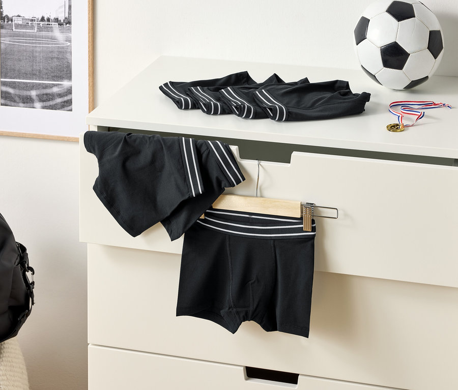 Sur une commode sont posés des boxers noirs à rayures blanches, un ballon de foot et une médaille. Un short de sport est suspendu à un cintre.