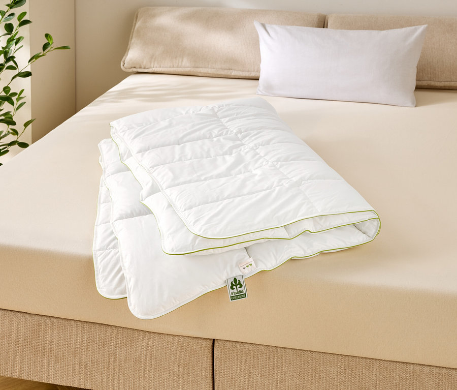 Une couette blanche pliée et matelassée avec une bordure verte sur un lit beige avec deux oreillers et une plante en arrière-plan.