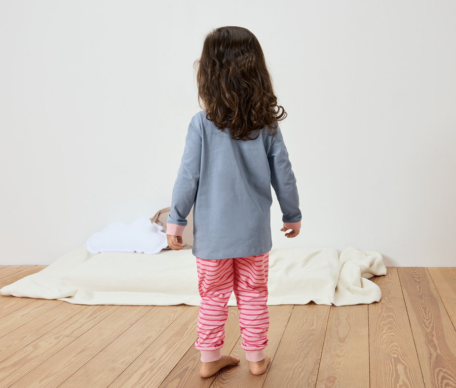 Une petite fille avec de longs cheveux bruns bouclés se tient dos au spectateur, portant un haut de pyjama bleu-gris et un pantalon de pyjama rayé rose, debout sur un plancher en bois.