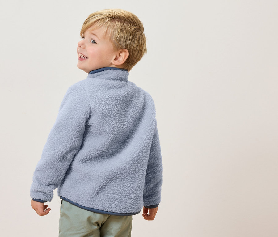 Un petit garçon aux cheveux blonds porte une veste polaire bleu clair et un pantalon vert clair, regardant sur le côté.