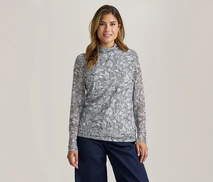 Une femme souriante aux cheveux bruns porte un haut à manches longues gris et blanc à motif peau de serpent avec un col montant et un pantalon large foncé.
