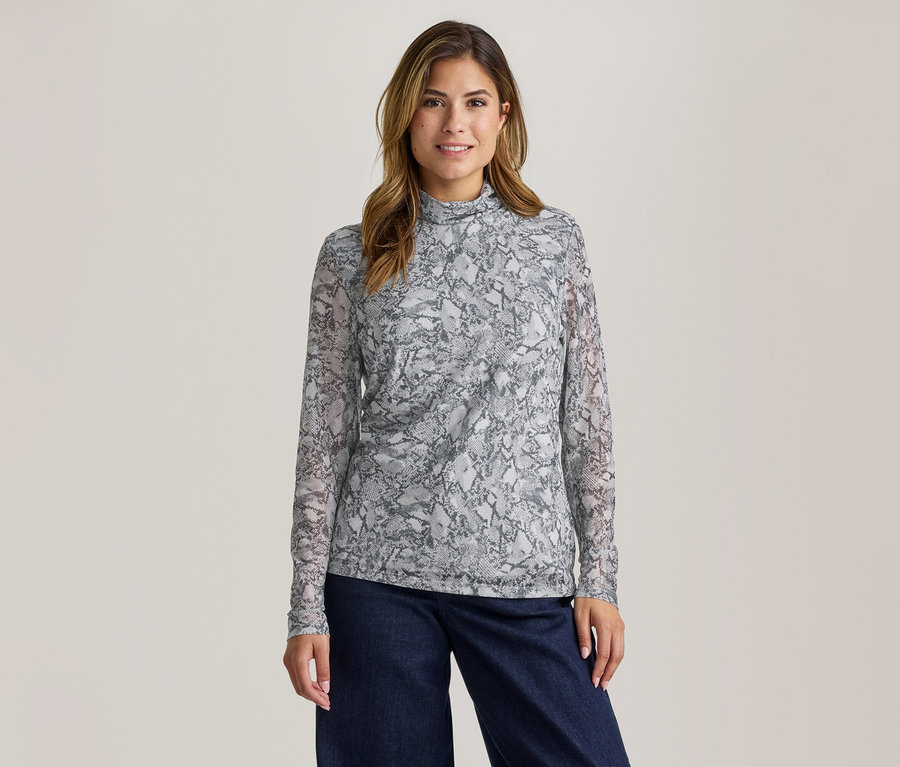 Une femme souriante aux cheveux bruns porte un haut à manches longues gris et blanc à motif peau de serpent avec un col montant et un pantalon large foncé.