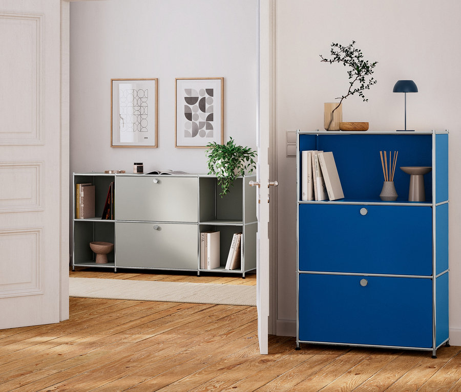 Une porte blanche ouverte relie deux pièces aux planchers en bois. Au premier plan, une unité de rangement modulaire bleue comporte une étagère supérieure ouverte avec des livres, un diffuseur, une lampe et une plante, ainsi que deux sections inférieures fermées. À l'arrière-plan, une unité de rangement modulaire gris clair avec des sections ouvertes et fermées contient des livres et une plante, avec deux œuvres d'art abstraites encadrées au-dessus.