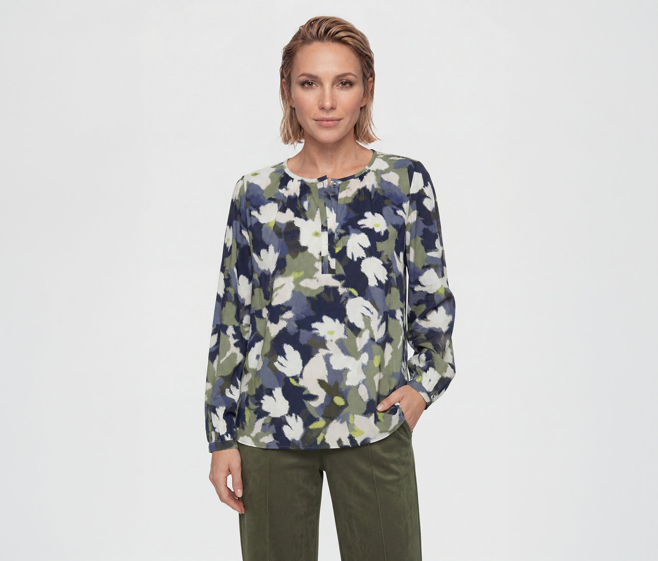Femme portant une blouse à manches longues avec un motif floral abstrait bleu, vert et blanc, associée à un pantalon vert olive. Sa main droite est dans sa poche.