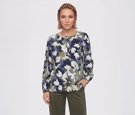 Femme portant une blouse à manches longues avec un motif floral abstrait bleu, vert et blanc, associée à un pantalon vert olive. Sa main droite est dans sa poche.