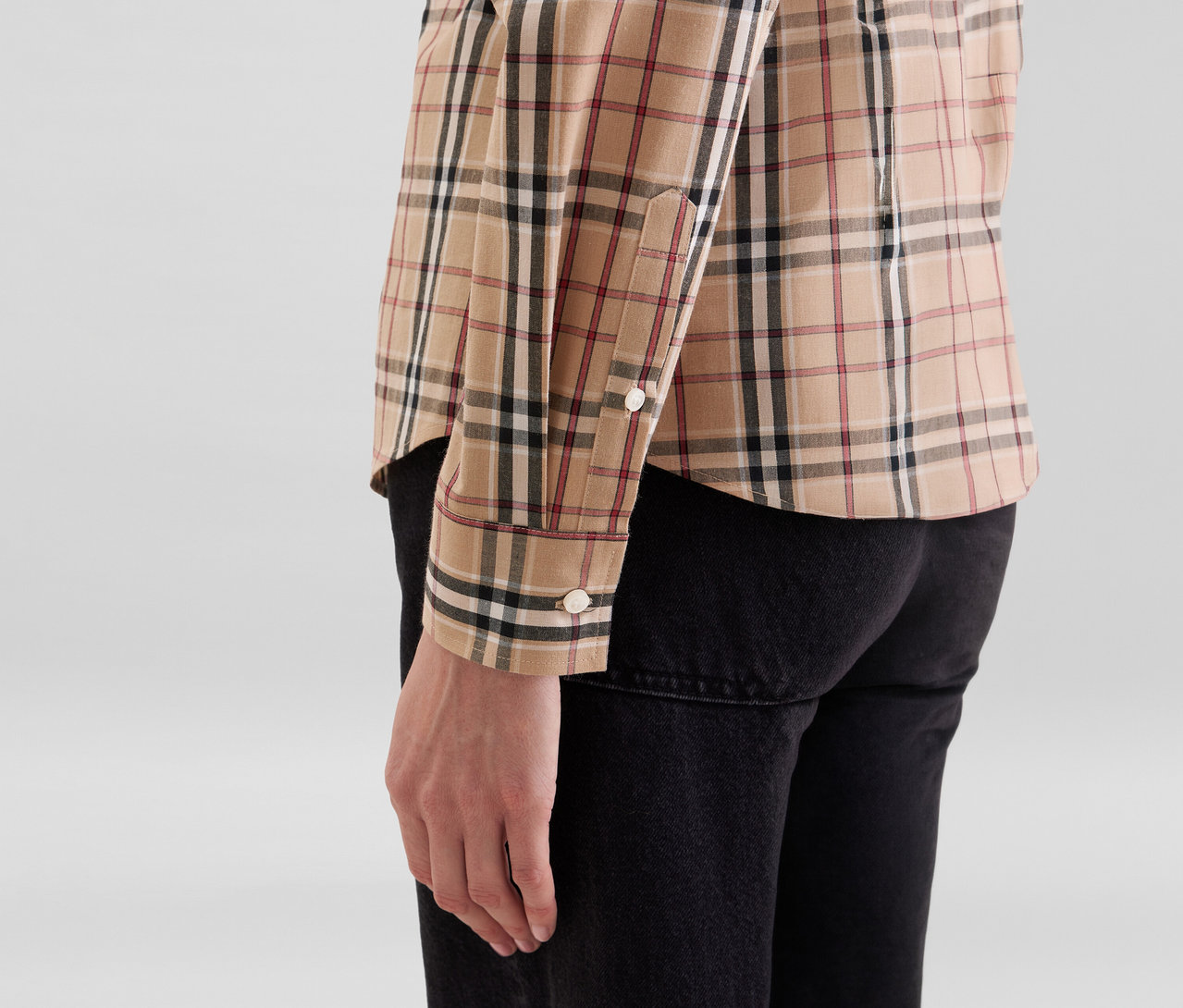 Personne portant une chemise à manches longues à carreaux beige avec des lignes noires, blanches et rouges, associée à un pantalon noir, vue de côté.