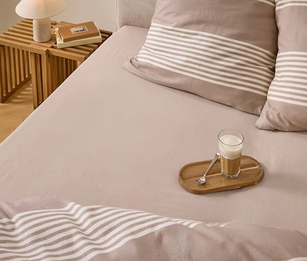 Un lit avec une literie marron clair, des oreillers et une couette rayés. Un verre de café latté sur un plateau en bois avec une cuillère est posé sur le lit. Une table de chevet en bois avec une lampe et des livres est à côté du lit.