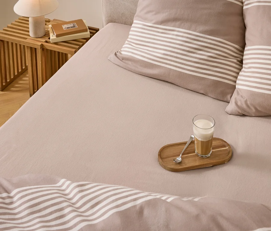 Un lit avec une literie marron clair, des oreillers et une couette rayés. Un verre de café latté sur un plateau en bois avec une cuillère est posé sur le lit. Une table de chevet en bois avec une lampe et des livres est à côté du lit.
