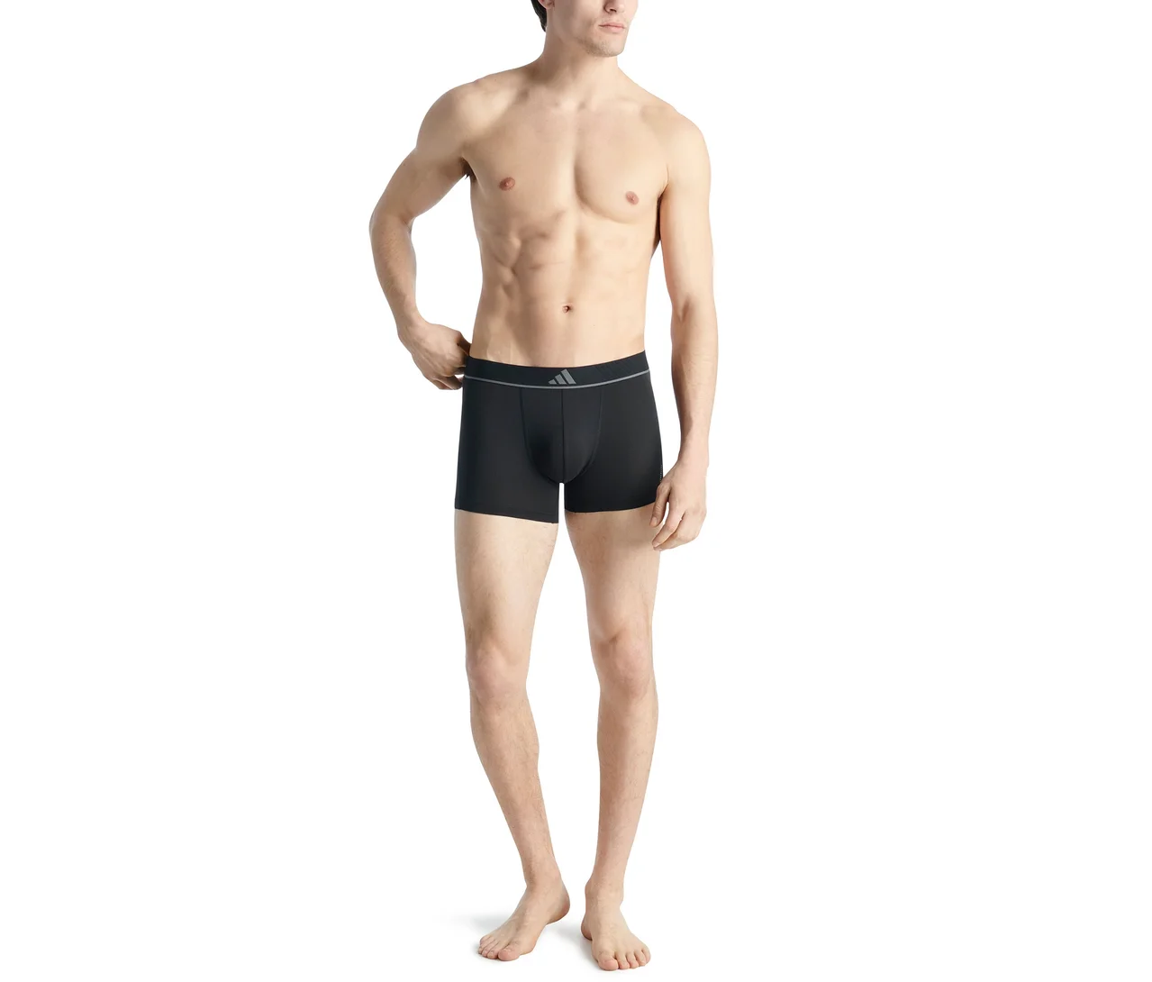 Un homme torse nu se tient debout, portant un boxer noir avec une ceinture grise affichant un logo Adidas, ajustant la ceinture de sa main droite.