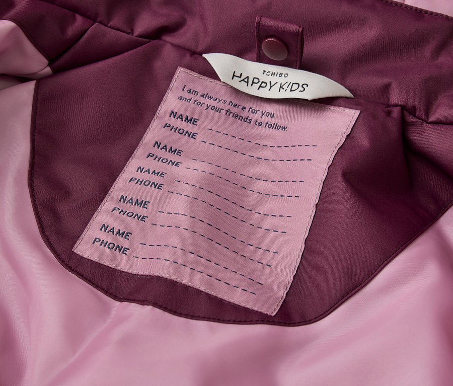 Gros plan sur une étiquette rose cousue à l'intérieur d'une veste violette et rose. L'étiquette porte l'inscription "TCHIBO HAPPY KIDS" en haut, suivie de "I am always here for you and for your friends to follow.". En dessous se trouvent cinq paires de lignes pour "NAME" et "PHONE".