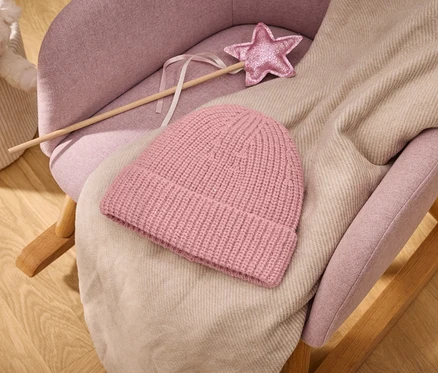 Bonnet tricoté rose et plaid beige clair sur un rocking-chair rembourré rose avec une baguette magique rose pailletée en forme d'étoile; à l'arrière-plan, un panier avec une peluche sur un plancher en bois.