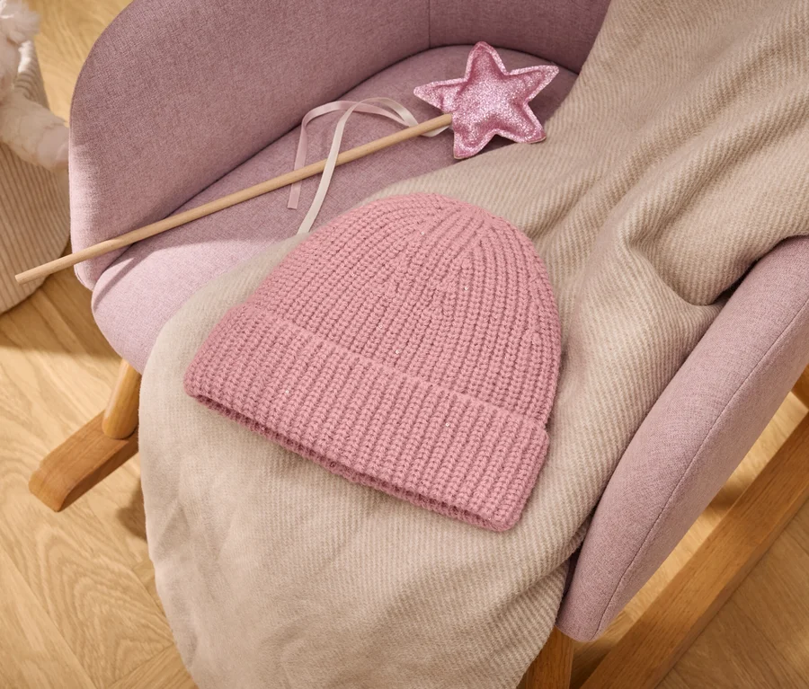 Bonnet tricoté rose et plaid beige clair sur un rocking-chair rembourré rose avec une baguette magique rose pailletée en forme d'étoile; à l'arrière-plan, un panier avec une peluche sur un plancher en bois.