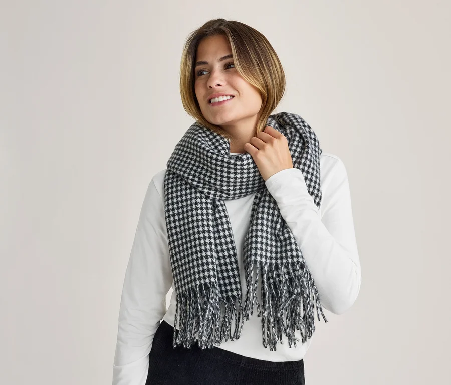 Une femme souriante portant une écharpe à motif pied-de-poule noir et blanc avec des franges, regardant sur le côté, une main près de l'écharpe.
