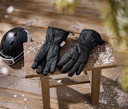 Casque de ski noir avec lunettes blanches et gants de ski noirs posés sur un tabouret en bois et un plancher en bois, parsemés de neige artificielle.