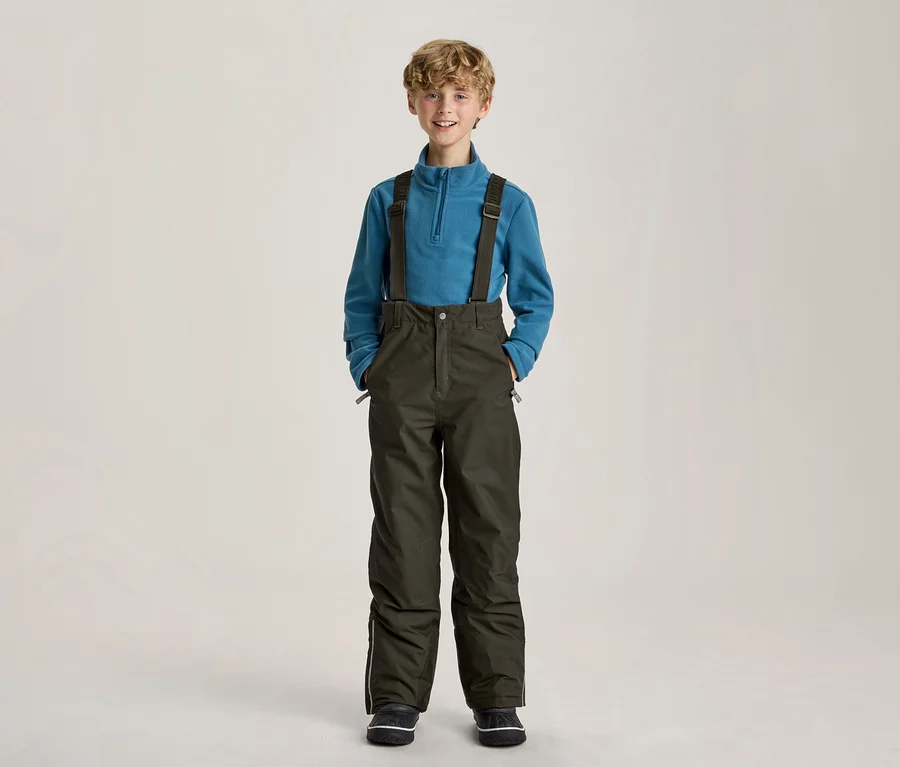 Jeune garçon souriant portant un pull polaire bleu et un pantalon de ski vert foncé à bretelles, les mains dans les poches.