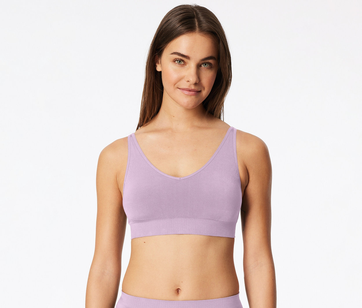 Femme aux longs cheveux bruns portant une bralette lilas à col en V et larges bretelles, regardant devant elle sur fond blanc.