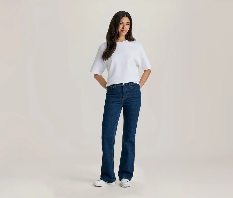 Une femme souriante aux longs cheveux foncés porte un t-shirt blanc, un jean évasé bleu foncé et des baskets blanches, les mains derrière le dos.