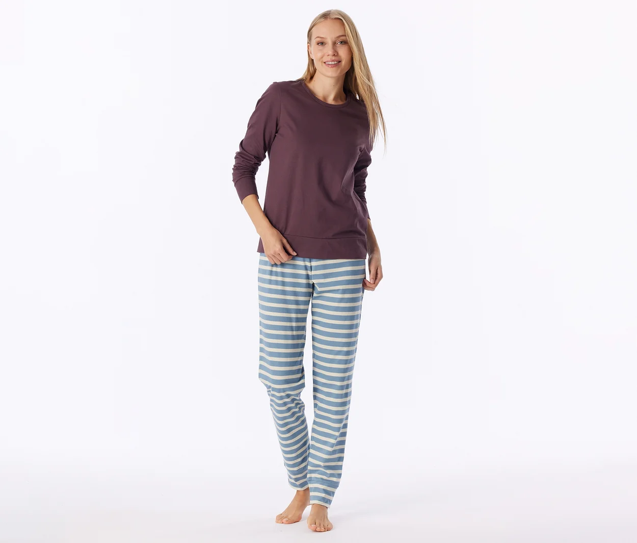 Une femme porte un haut à manches longues violet foncé et un pantalon de pyjama bleu clair à rayures horizontales crème. Elle a de longs cheveux blonds, sourit et est pieds nus.