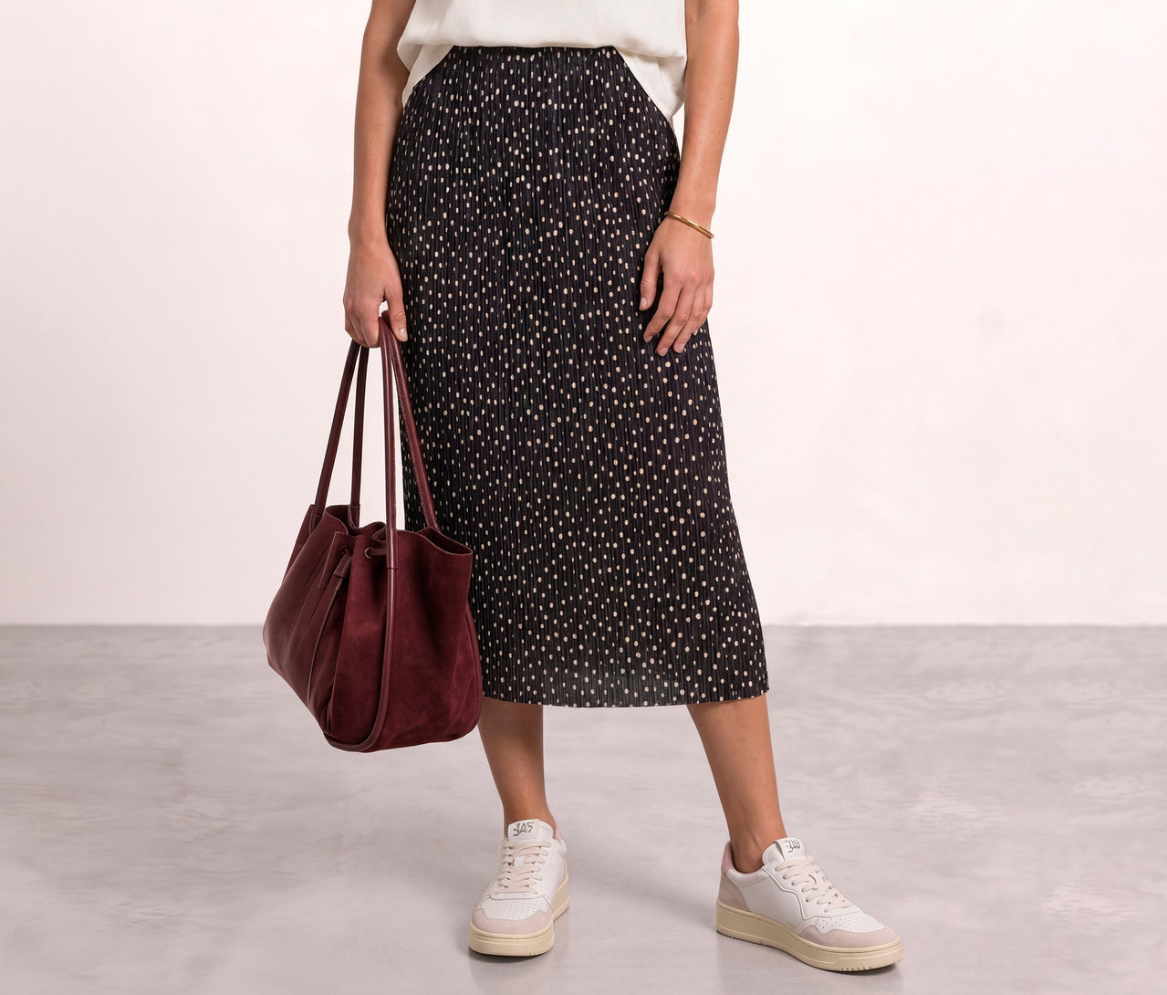 Personne portant un haut blanc et une jupe midi Street One noire à pois blancs, tenant un sac à main en daim bordeaux et portant des baskets blanches.