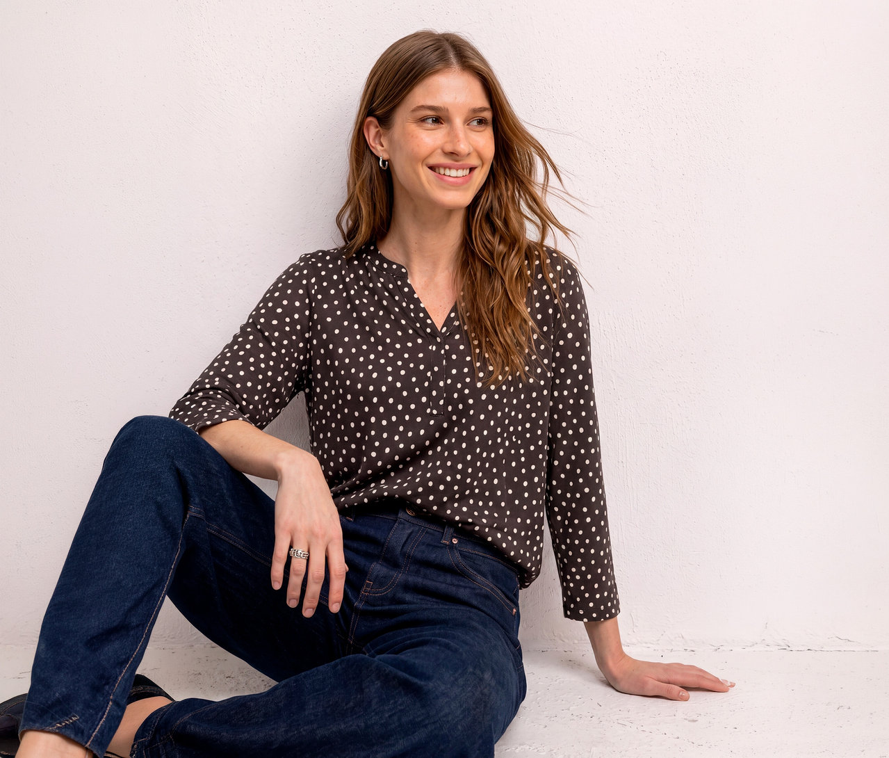 Femme assise souriante contre un mur blanc, portant un T-shirt chemisier Street One marron foncé à pois blancs et un jean bleu foncé.
