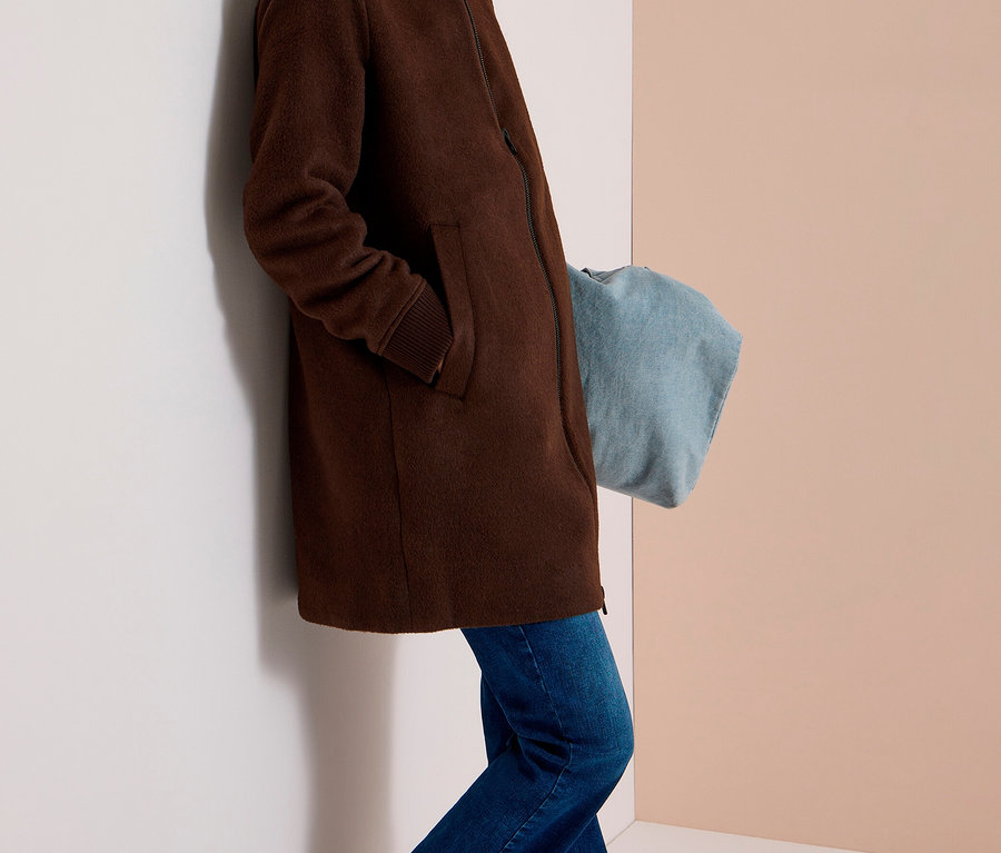 Femme souriante portant un blouson CECIL marron, un jean bleu et des baskets argentées, appuyée contre un mur.
