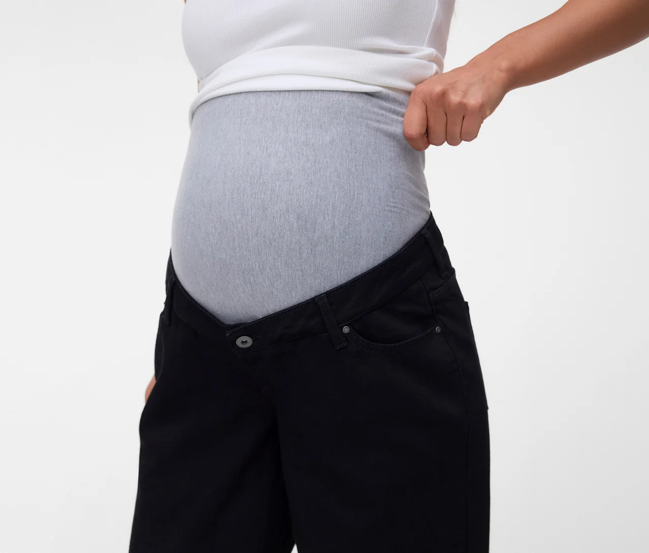 Torse de femme enceinte portant un débardeur blanc et un jean de maternité noir avec un empiècement élastique gris, sa main remontant le débardeur.