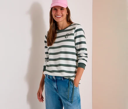 Femme souriante portant une casquette rose, un sweatshirt CECIL rayé blanc et vert avec un logo « C », et un jean bleu clair, la main dans la poche.