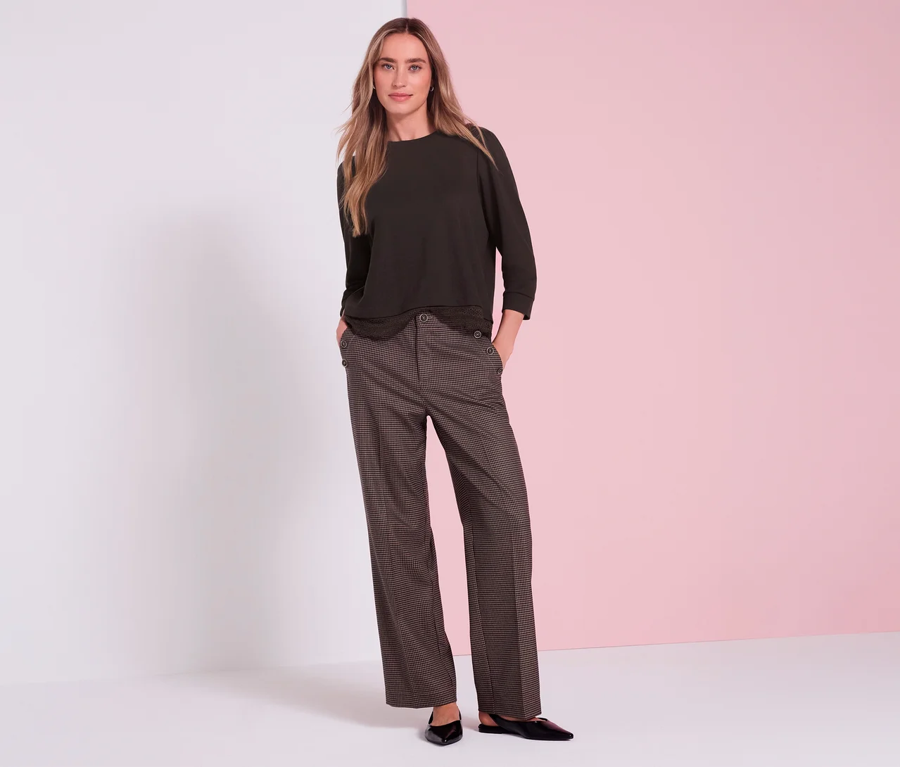 Une femme aux longs cheveux blonds, les mains dans les poches, porte un T-shirt en jersey 3/4 Street One marron, un pantalon large marron à motifs avec des boutons décoratifs sur les poches et des chaussures plates noires, devant un fond blanc et rose.