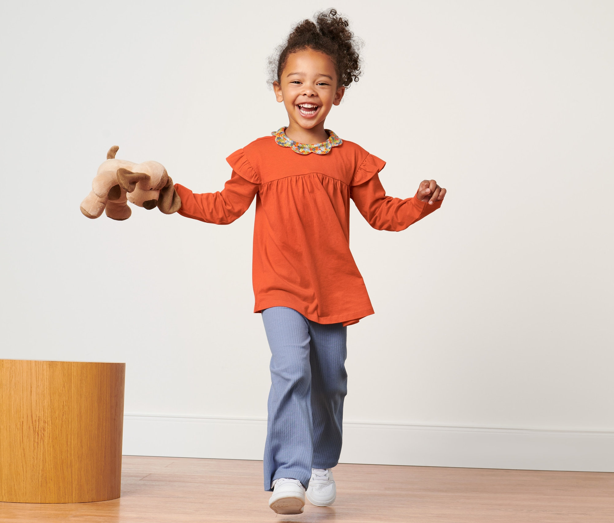 Fillette souriante portant un t-shirt à manches longues orange, tenant un chien en peluche et marchant sur un plancher en bois.