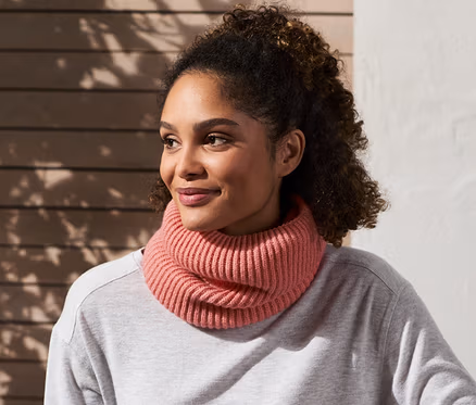 Une femme est assise sur un banc et porte une écharpe tube en maille rose.