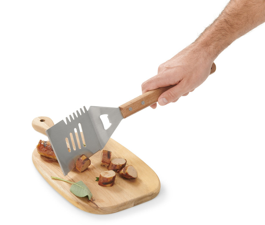 Une main tient un outil polyvalent pour barbecue au-dessus d'une planche à découper en bois avec des saucisses coupées.