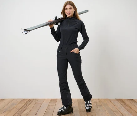 Une femme pose en pantalon de ski noir et chaussures de ski, avec des skis sur l'épaule.