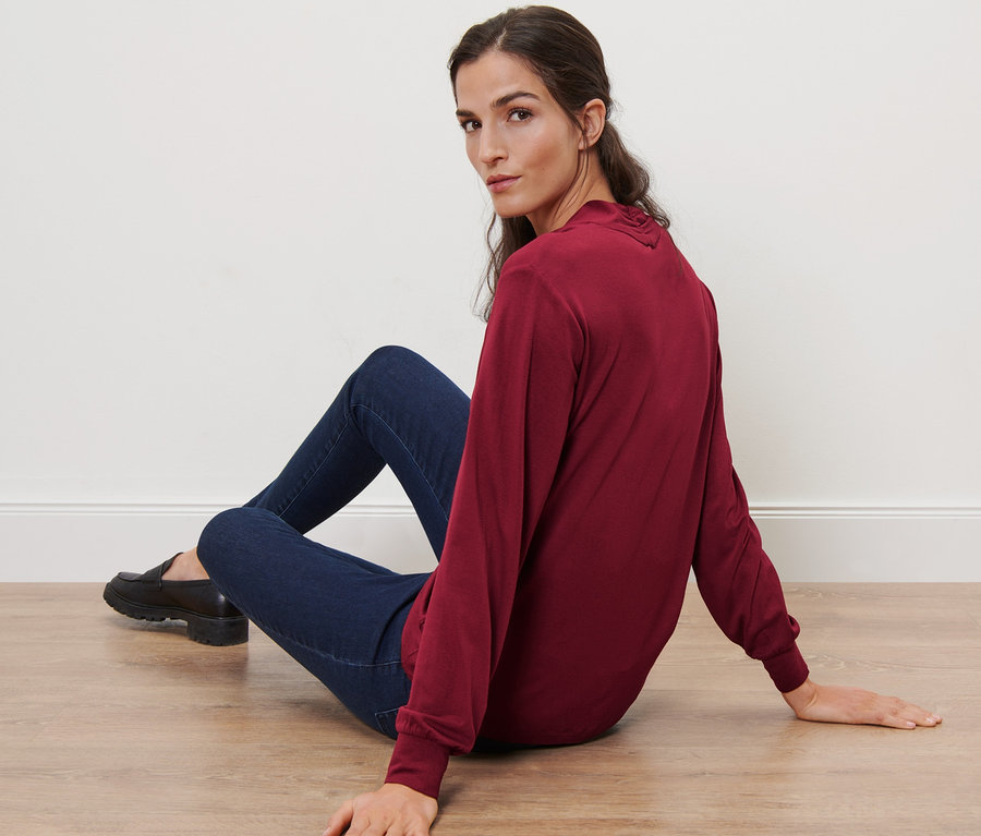 Une femme est assise par terre et porte un t-shirt manches longues avec encolure en V, rouge bordeaux.