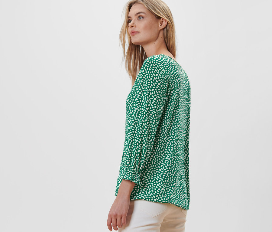 Une femme blonde pose de profil et porte une blouse imprimée verte.