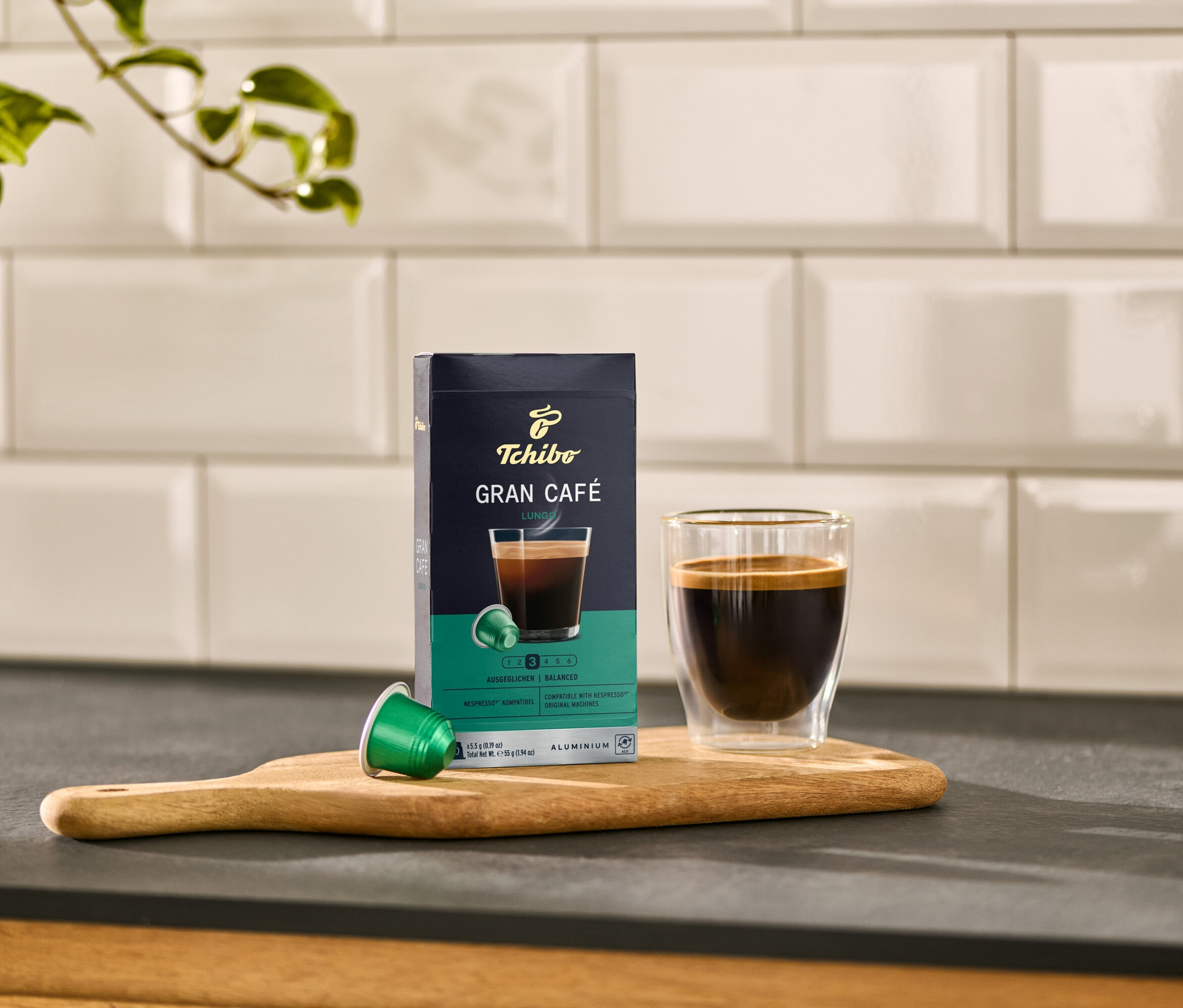 Sur une planche en bois, une boîte de Gran Café Lungo, une capsule verte et un verre de café.