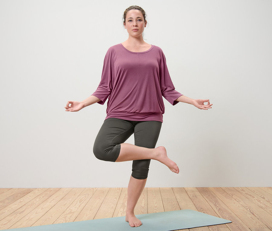 Femme debout sur un tapis de yoga dans la pose de l'arbre. Elle porte un t-shirt de yoga mauve et un collant de sport 3/4 olive.