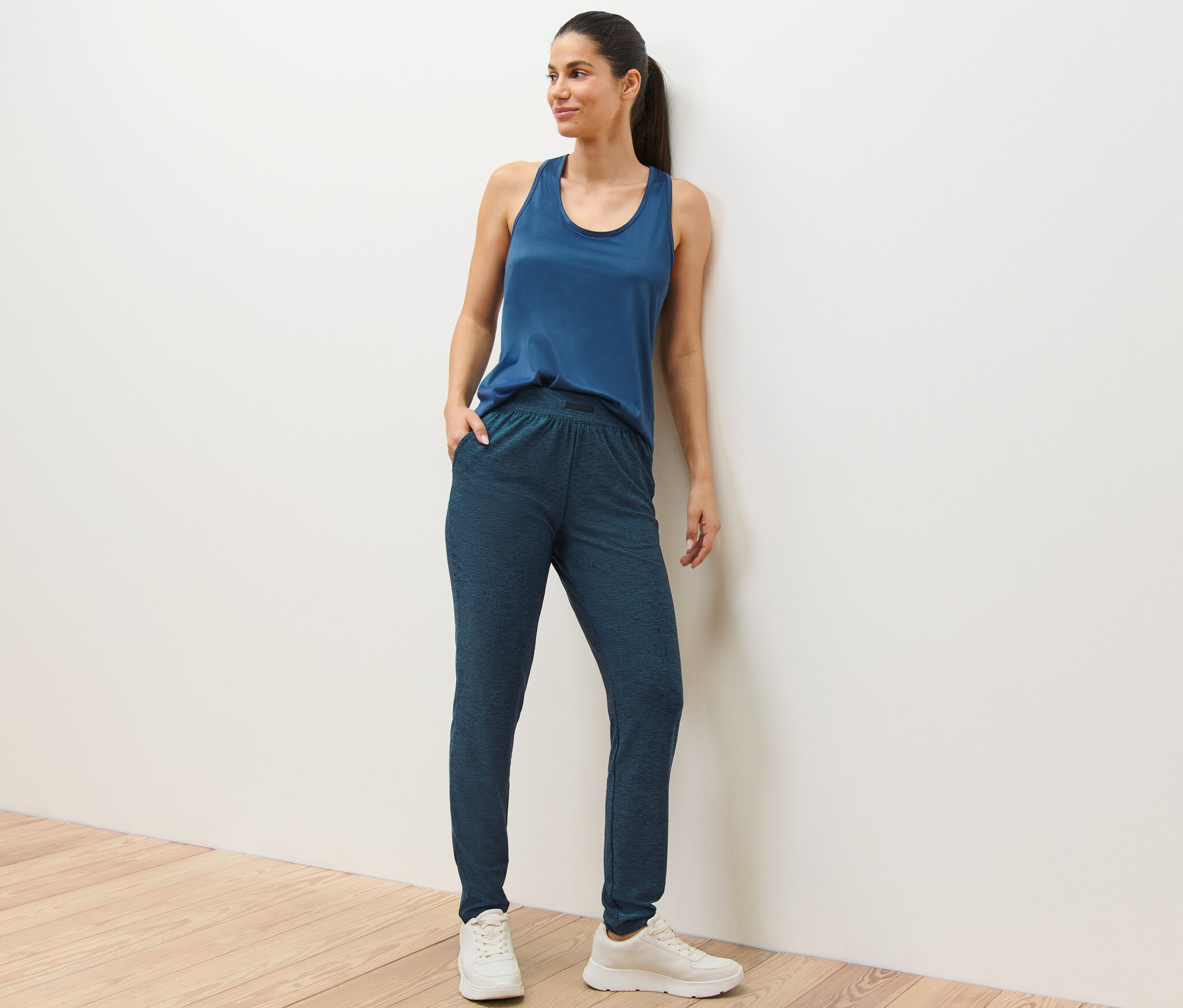 Une femme pose en pantalon de sport bleu et débardeur contre un mur blanc.