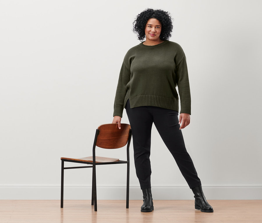 Une femme pose à côté d'une chaise, portant un pantalon stretch noir et un pull en tricot avec fente à la base.