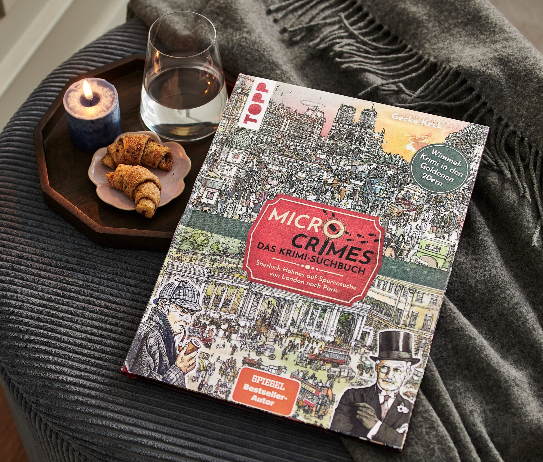 Sur un pouf se trouvent le livre « Micro Crimes – das Krimi-Suchbuch » (en Allemand), un verre d'eau, une bougie et une assiette de croissants.