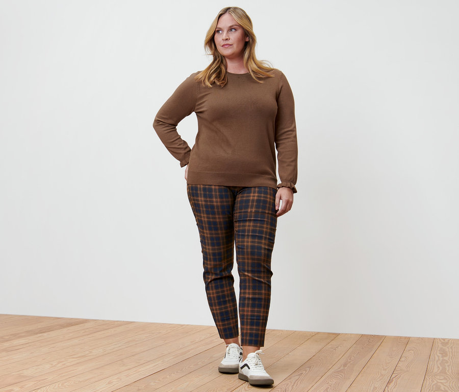 Femme posant dans un pull en maille fine avec finition ruchée, un pantalon stretch à carreaux et des baskets en cuir.