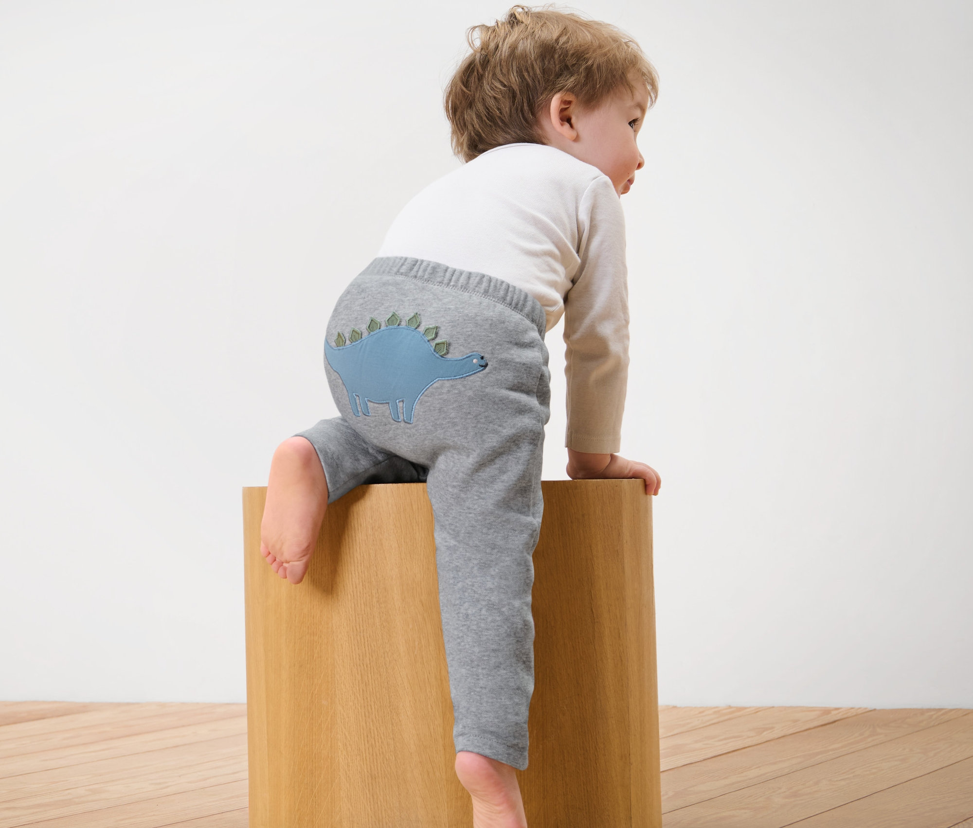 Un enfant est assis sur un cube en bois et porte un pantalon de survêtement gris avec un dinosaure.