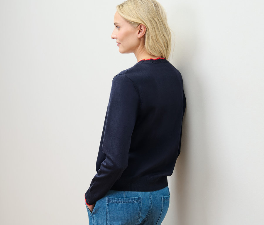 Une femme blonde se tient de côté et porte un cardigan bleu à maille fine avec bord ondulé et un jean.