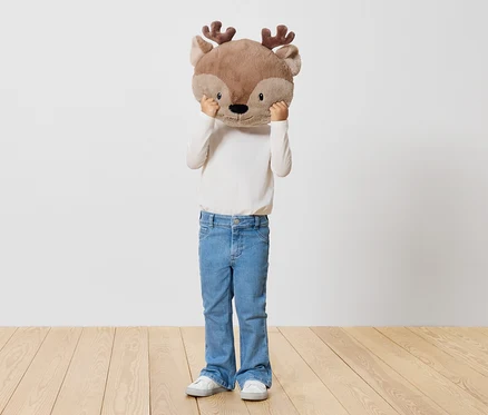 Un enfant tient une peluche de renne devant son visage, debout sur un plancher en bois devant un mur blanc.