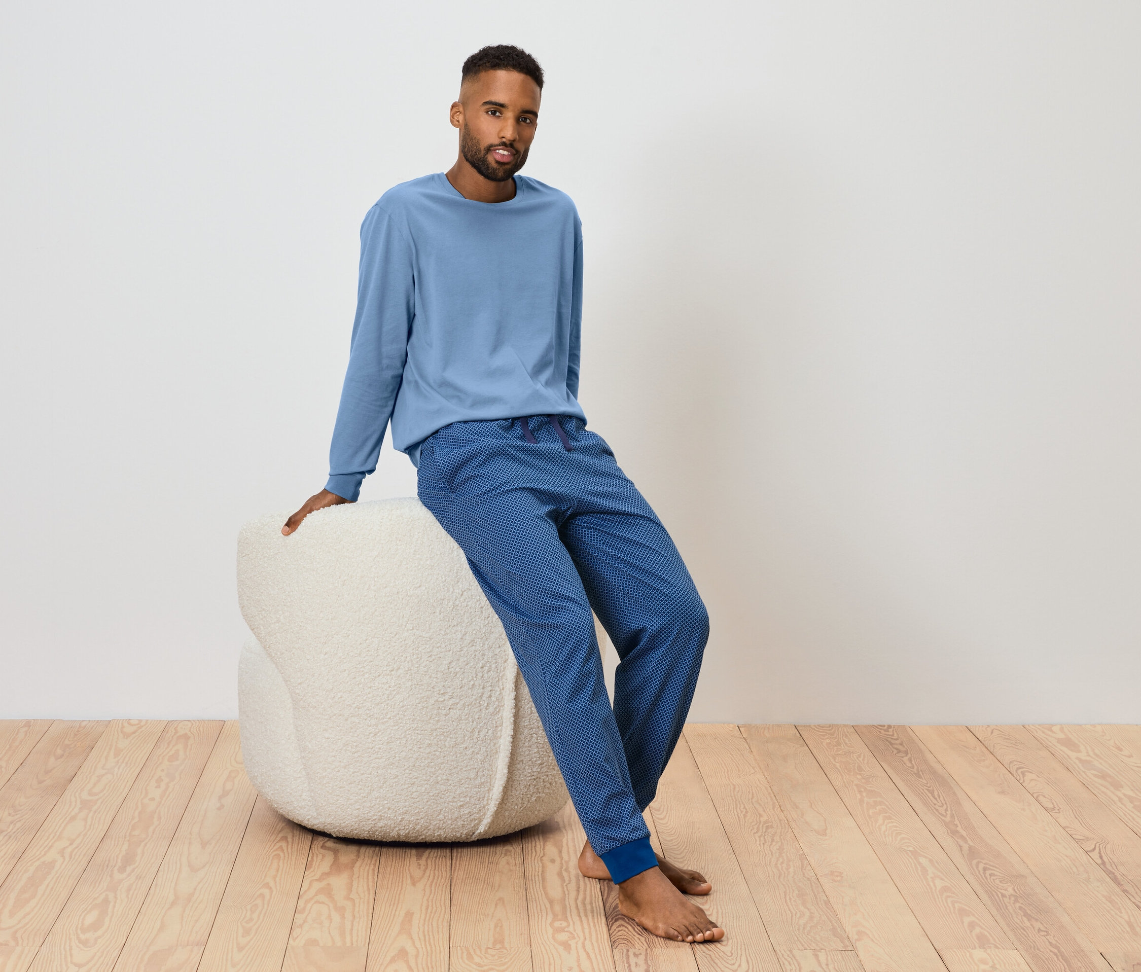 Un homme est assis sur un pouf blanc et porte un t-shirt bleu à manches longues et un pantalon de survêtement bleu.